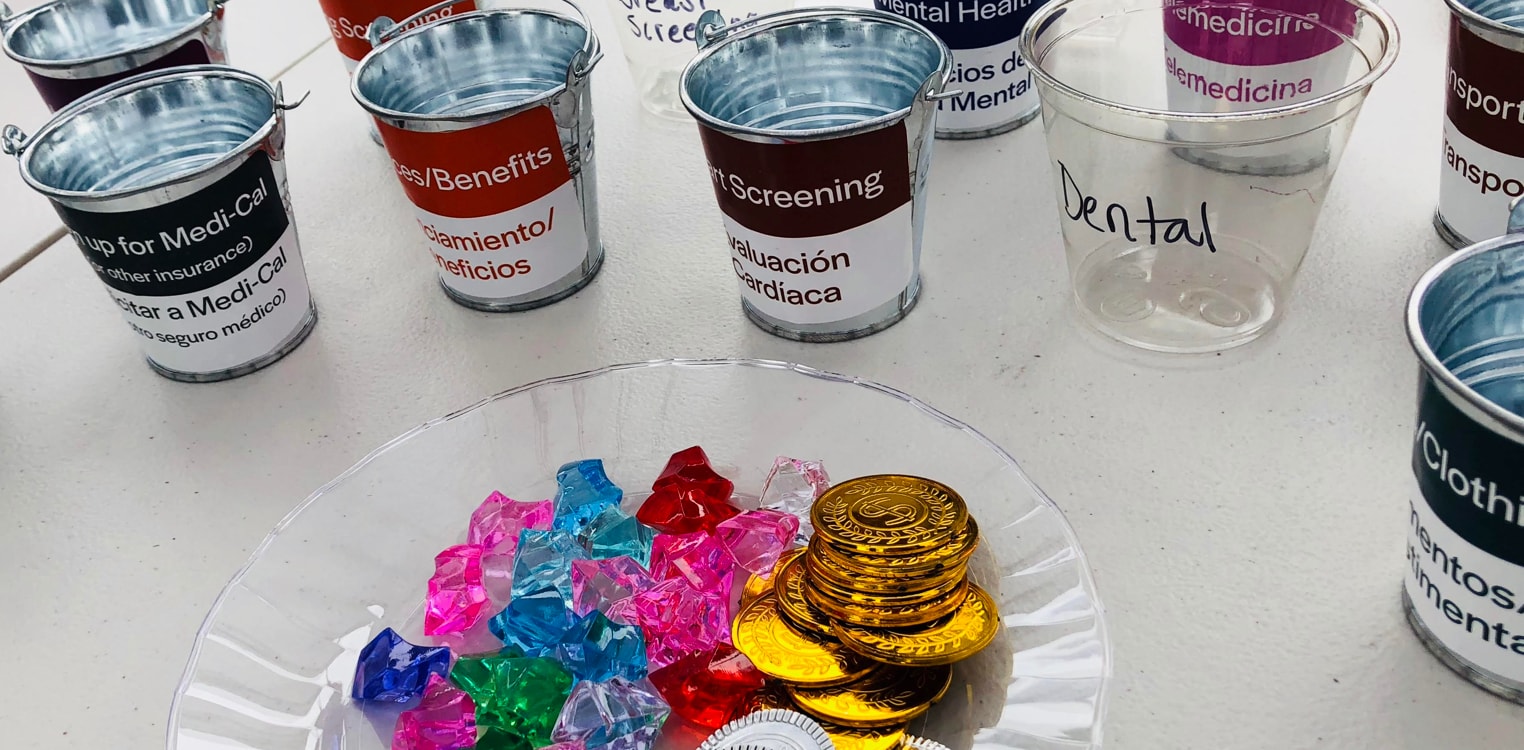Piles of toy jewels, gold and silver coins sit near empty cans labeled with health-related needs such as mental health, signing up for Medi-Cal, and health screening