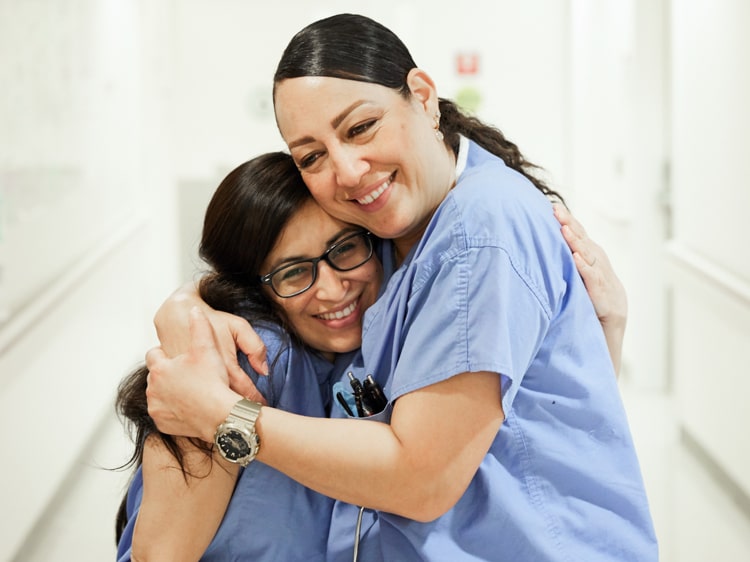 Two female nurses wearing blue scrubs smiling and hugging