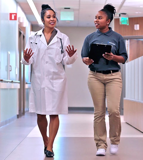 Mujer de color con bata médica blanca caminando por un pasillo hablando con una mujer de color joven