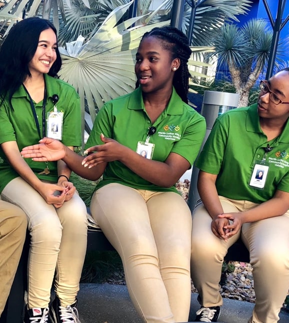 Three young female COPE scholars wearing green shirts talking in Healing Garden