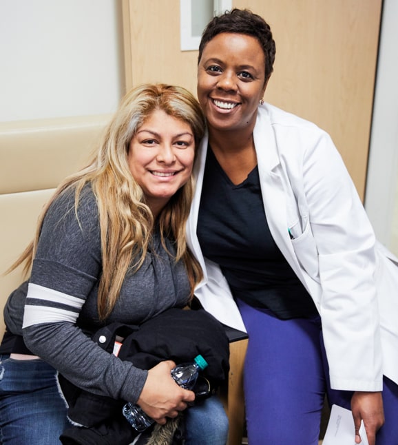 Una enfermera de color sentada junto a una paciente, ambas sonriendo
