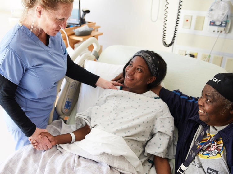 White nurse holds hand and smiles at Black female patient lying in hospital bed
