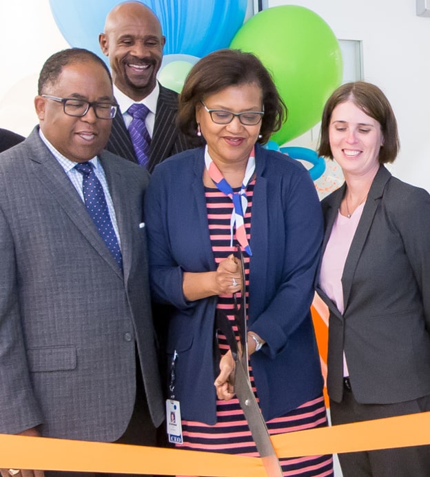 Dr. Batchlor, a middle-aged Black woman, cutting a large ceremonial ribbon with people gathered around her