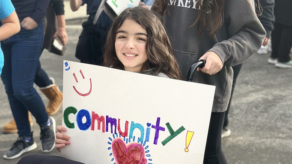 Photo of young girl in wheelchair holding a handmade sign that says "Community!"