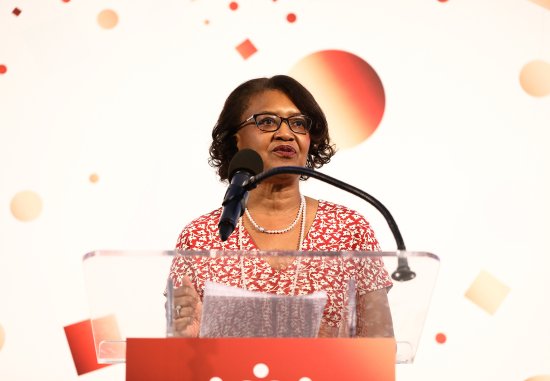 Dr. Elaine Batchlor, a middle-aged Black woman, speaking at a podium