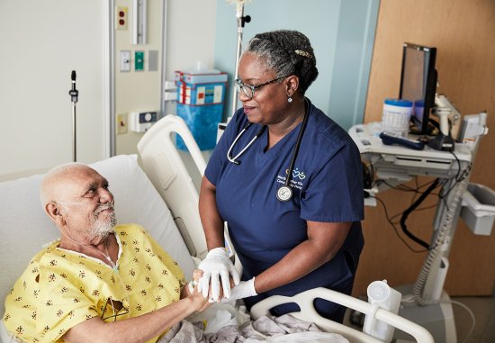 Black female nurse holding hands with older Latino patient in hospital bed