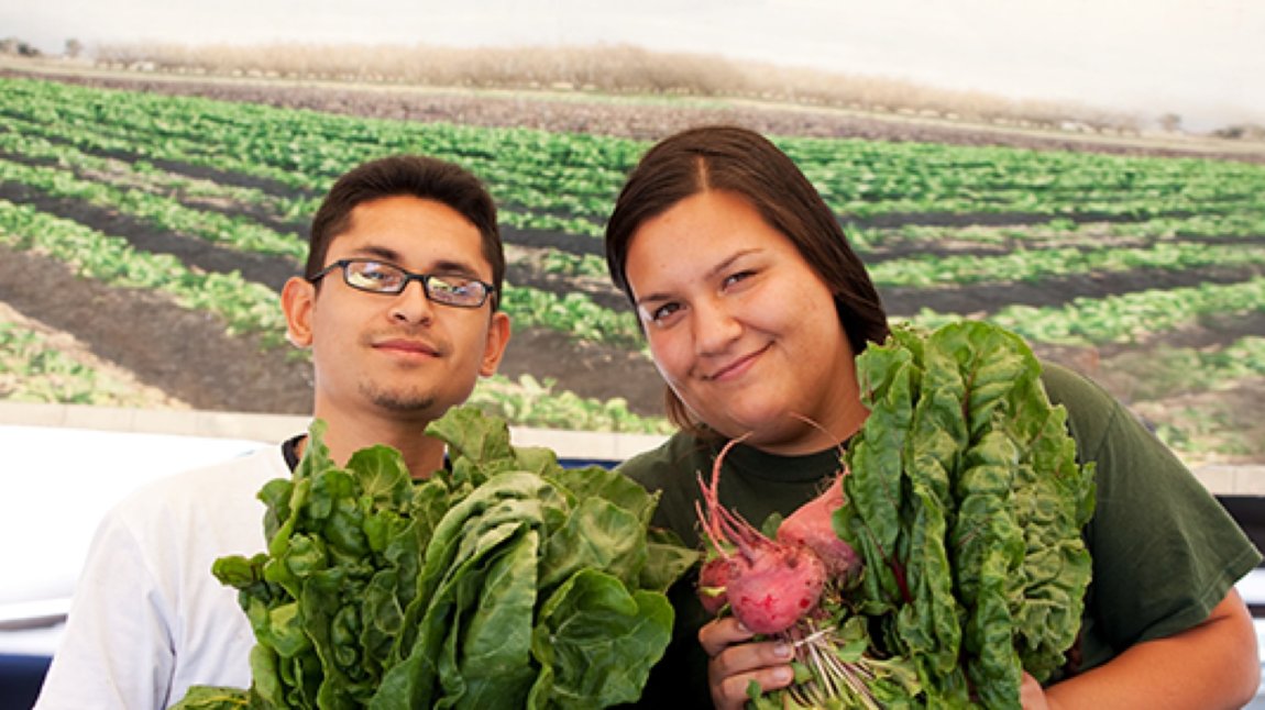 Young Latinx man and woman holding up leafy greens at farmers market