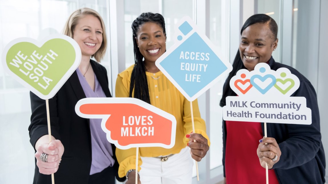 Three female staff members holding signs that say we love south LA, loves MLKCH, access equity life, and mlk community health foundation 