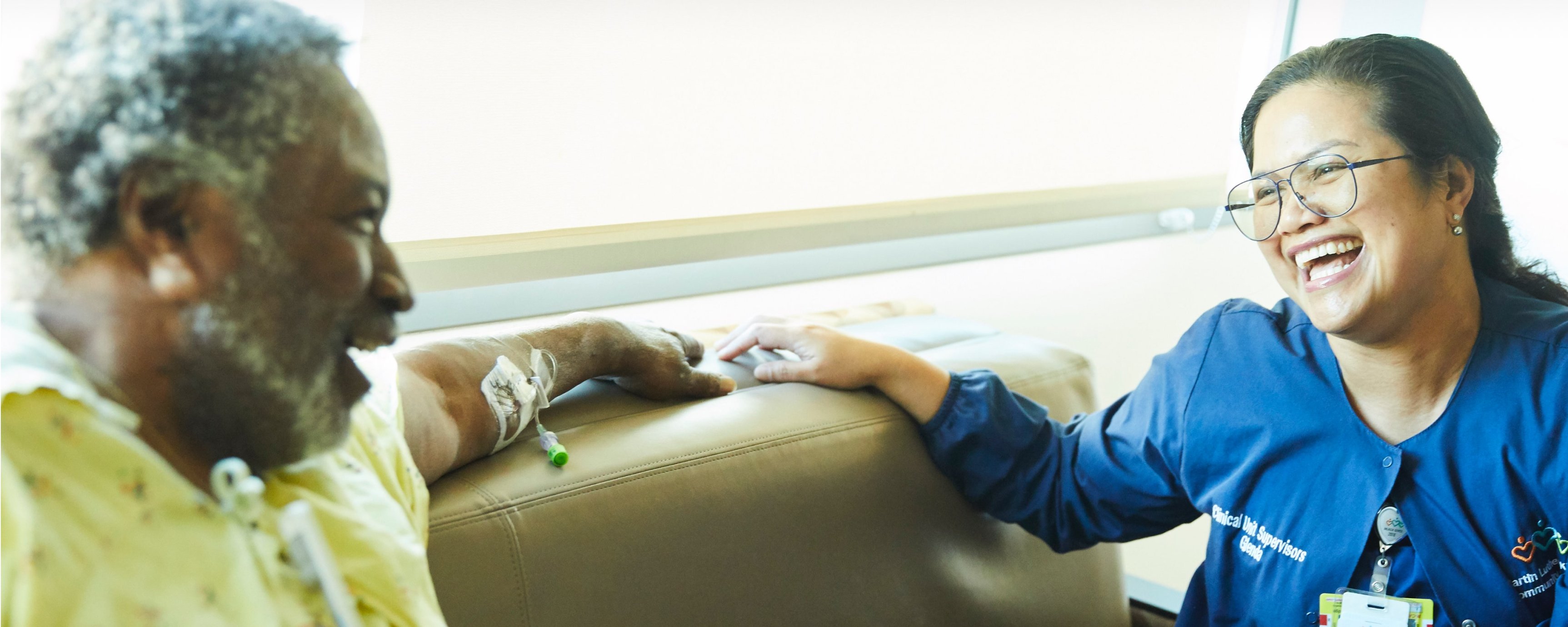 Older Black male patient laughing with young Asian female nurse on a couch