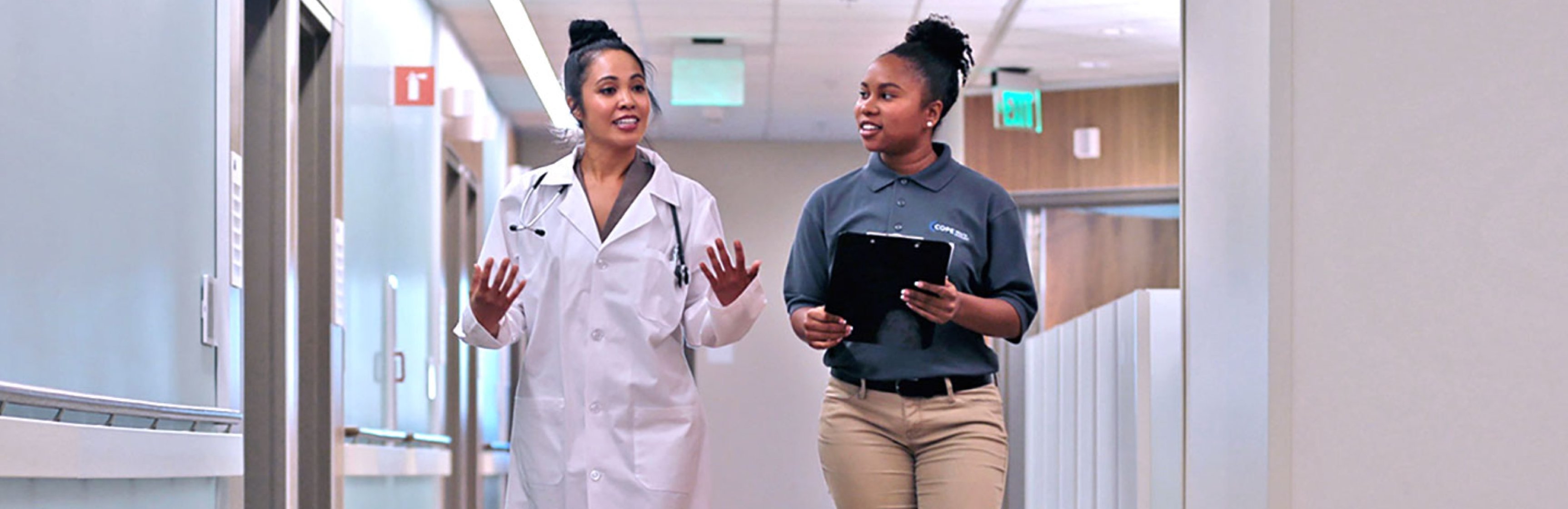 Black female in white doctors coat walking down a hall speaking to a young Black female