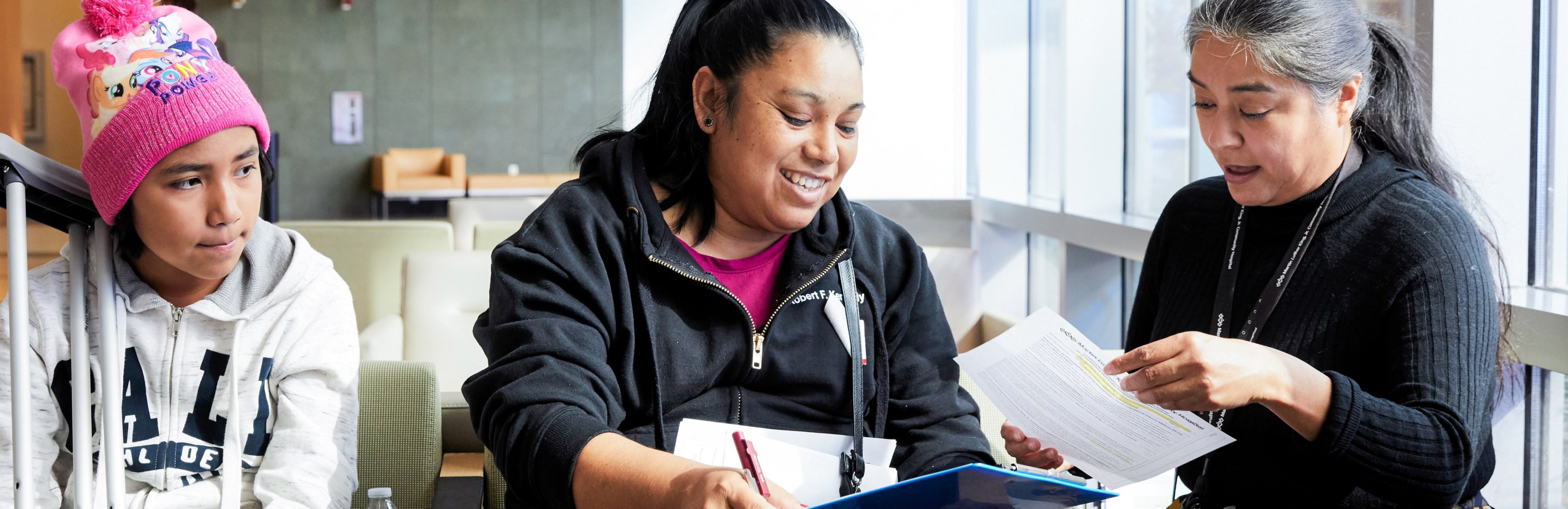 A female hospital employee reviews paperwork with a Latina woman in the waiting area