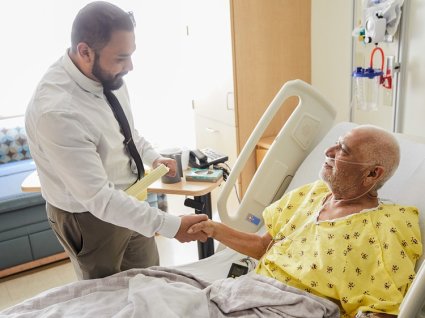 Male hospital administrator leaning down to shake the hand of older male patient lying in hospital bed