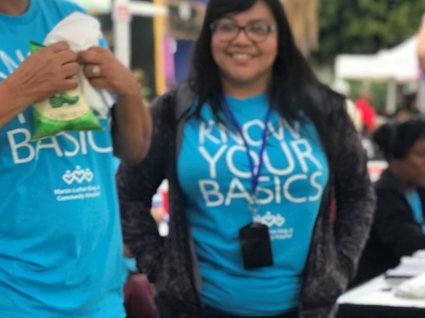 Mujer latina sonriendo y usando una camiseta de color azul brillante en la que se lee Conozca Lo Básico