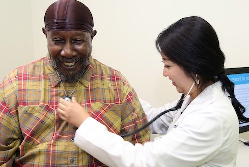 Female Asian doctor using stethoscope on smiling Black male patient's chest