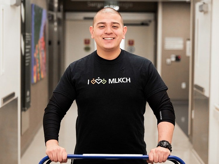 Smiling Latino staff member pushing a wheelchair down hospital hallway