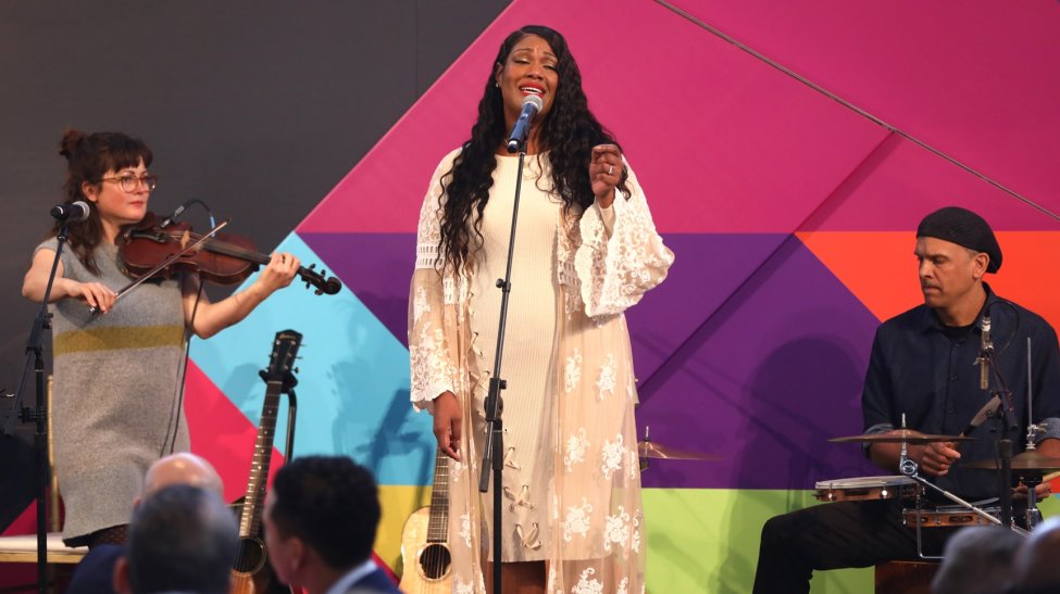 Keisha Renee, a young Black woman, singing with two white musicians on stage at the Dream Lunch